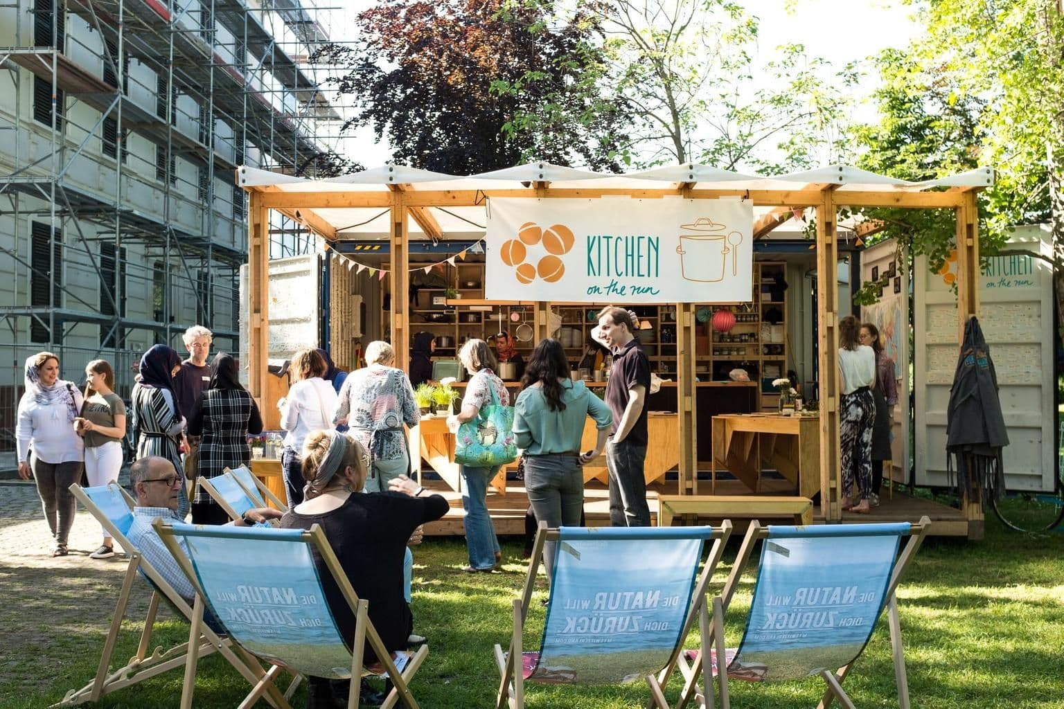 Ein Pavillon aus Holz lädt zum Verweilen ein. Viele Menschen stehen und sitzen drum herum.