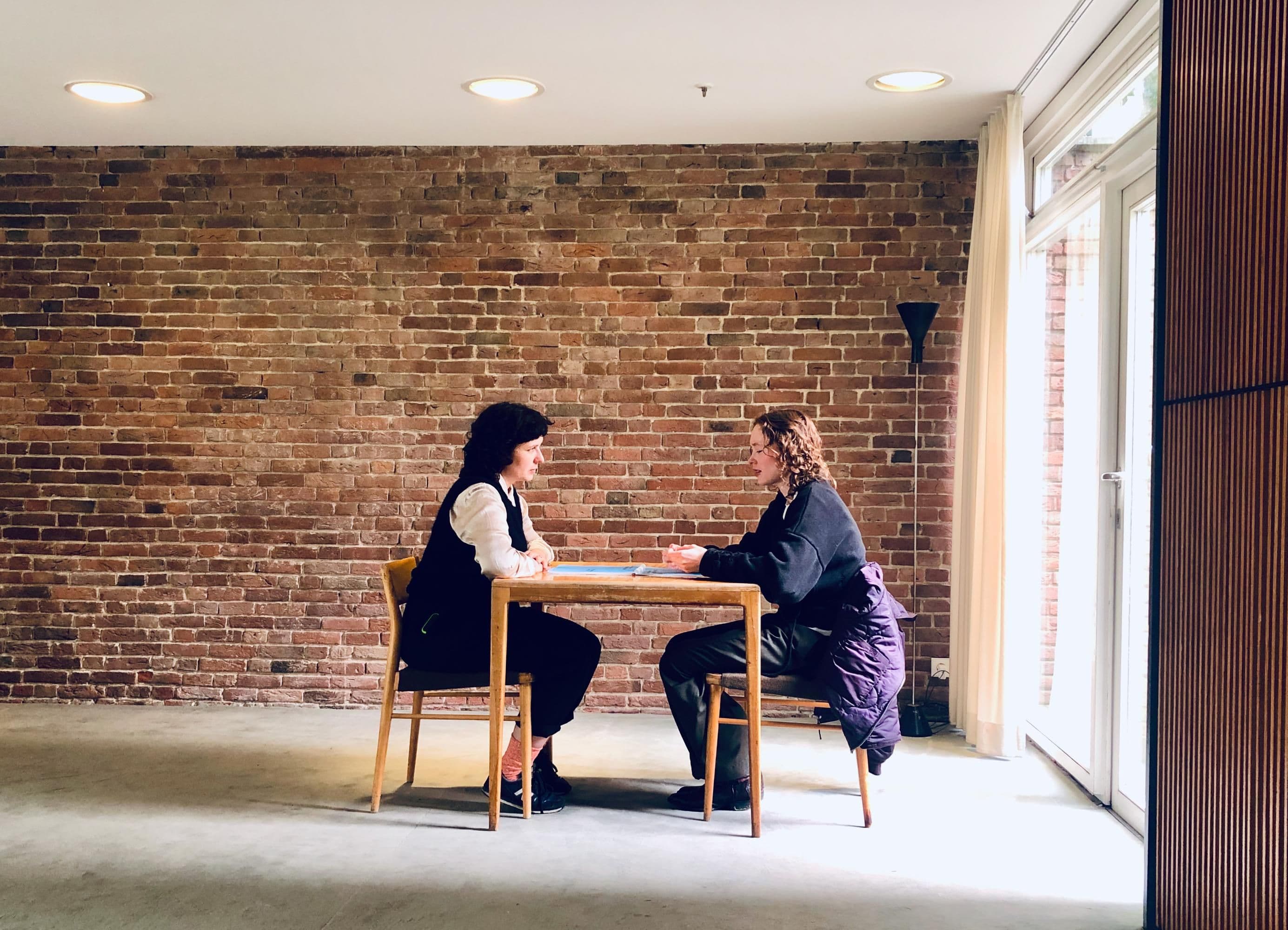 Zwei Personen sitzen an einem Tisch in einem offenen Raum.