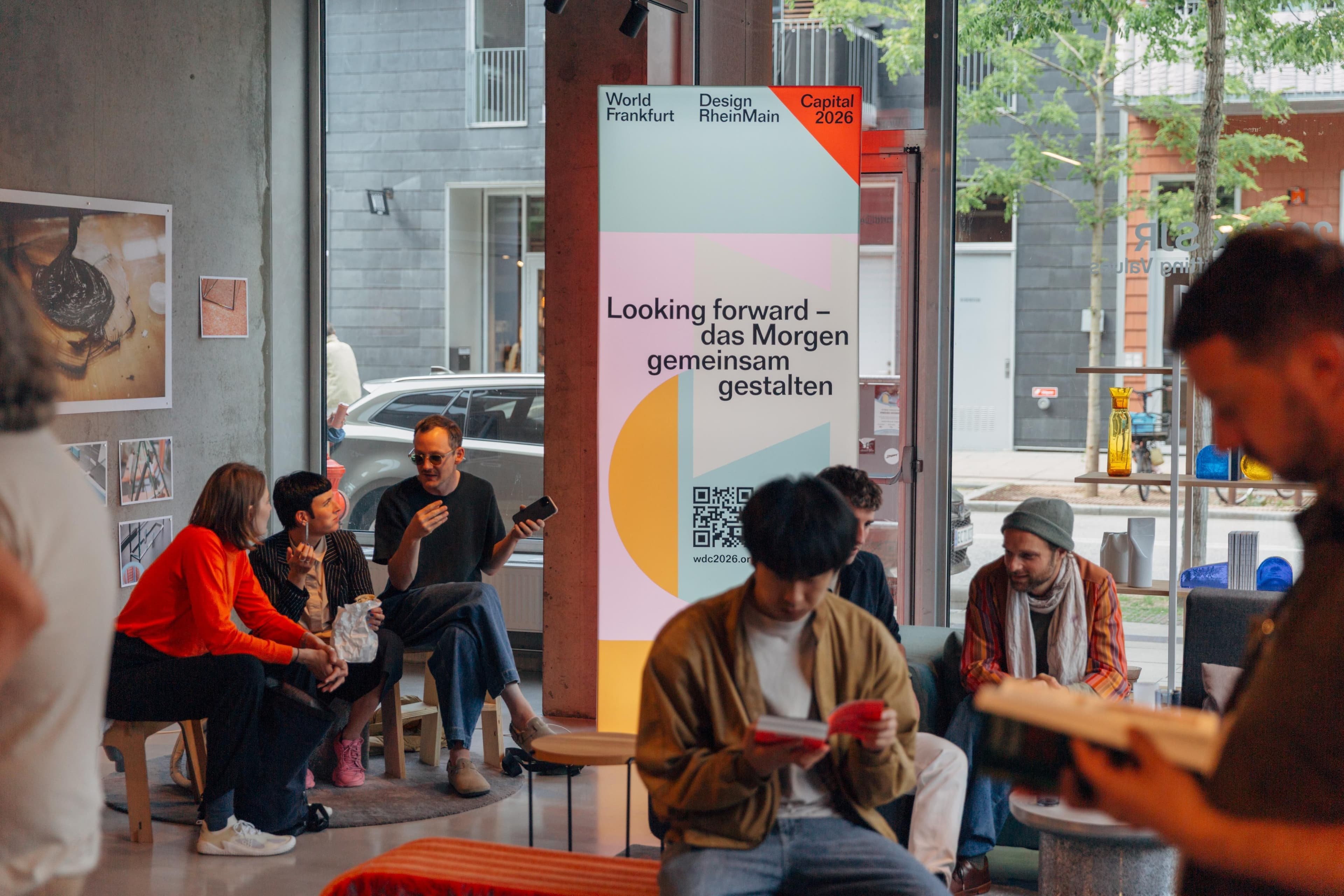 Einige Personen sitzen in einem schönen Raum mit viel Fensterfläche. Im Hintergrund ist ein großes Plakat von World Design Capital 2026.