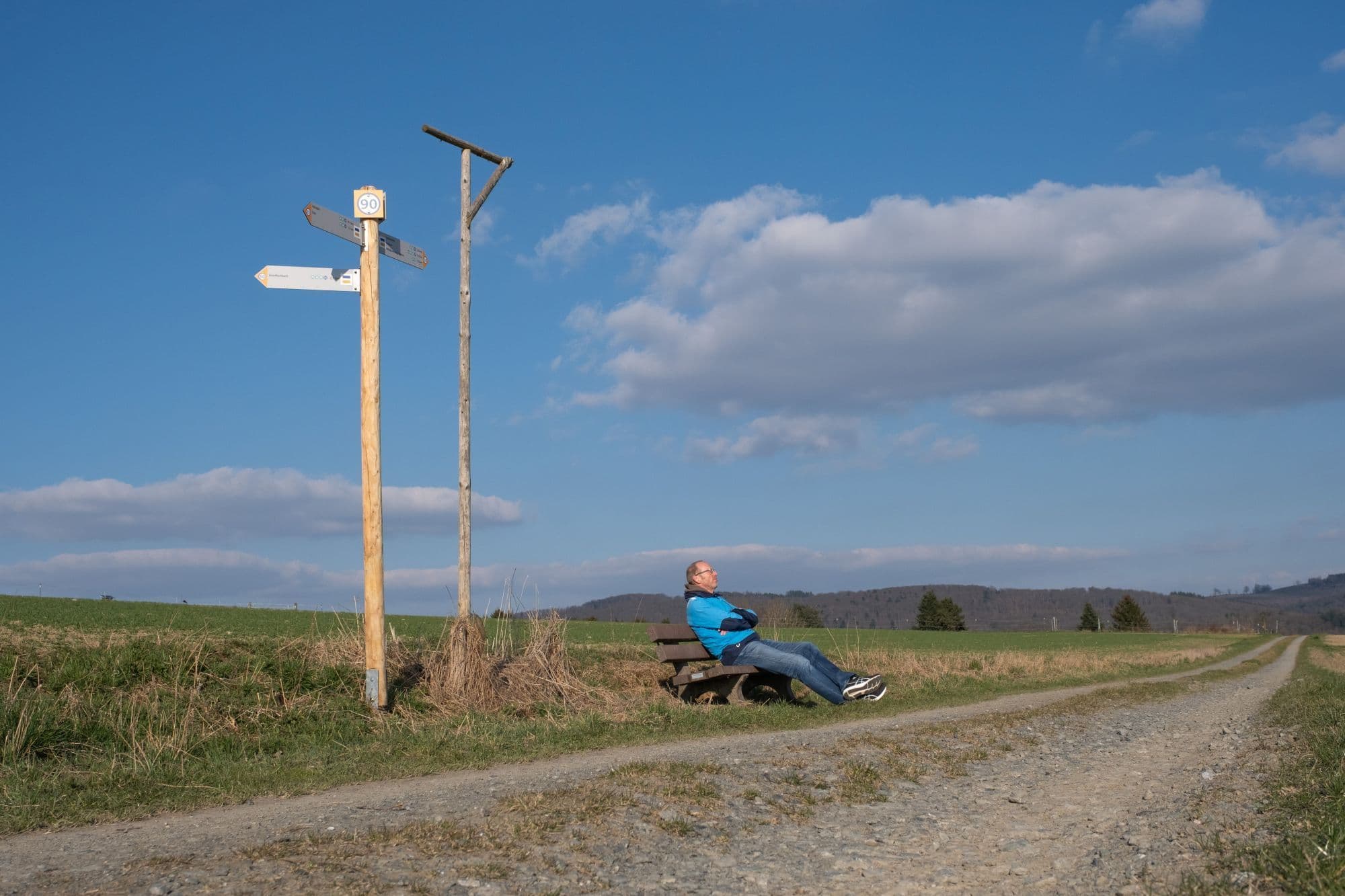 Auf einer Bank, die sich auf einer Wiese an einem Feldweg befindet, sitzt ein Mann.