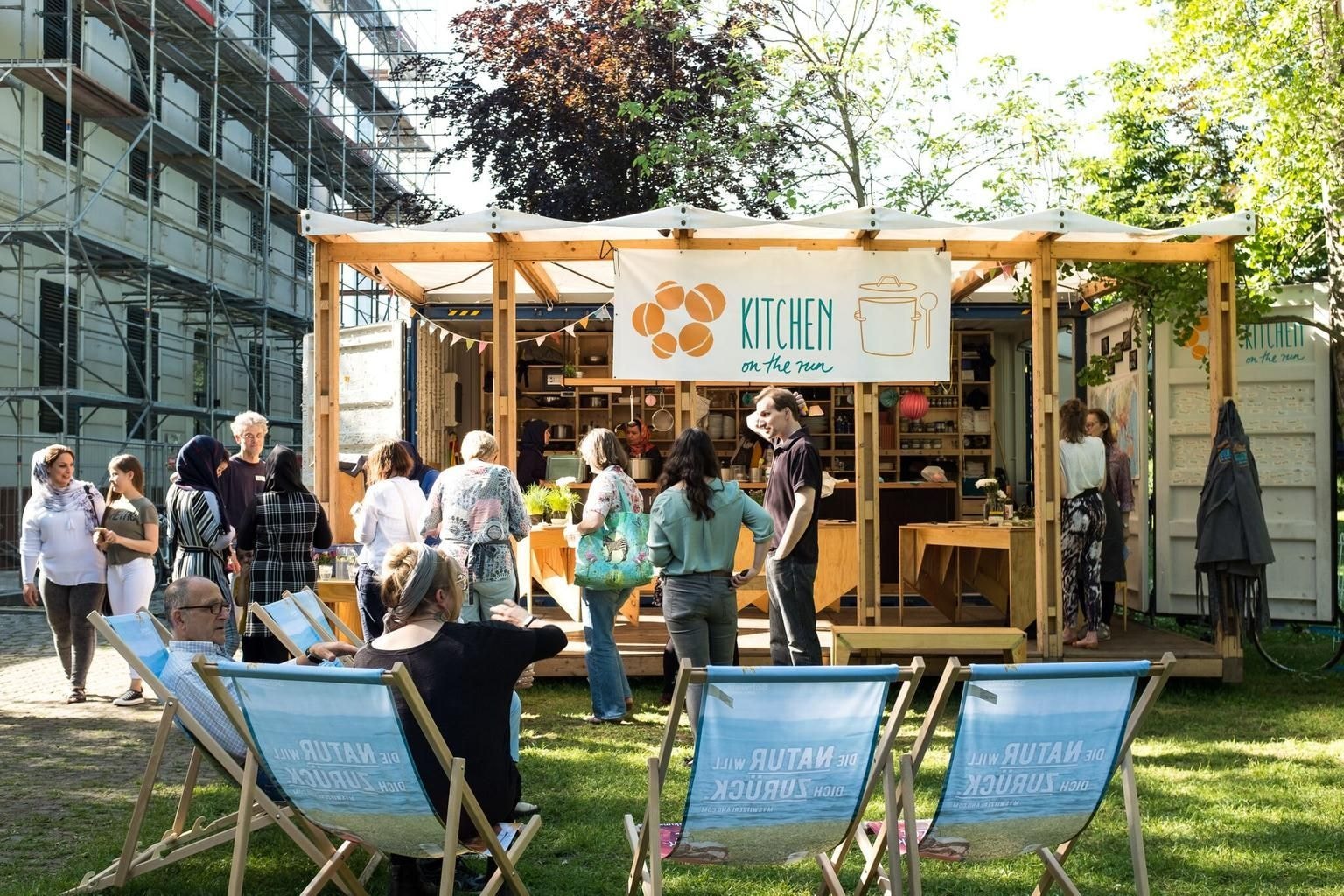 Ein Pavillon aus Holz lädt zum Verweilen ein. Viele Menschen stehen und sitzen drum herum.