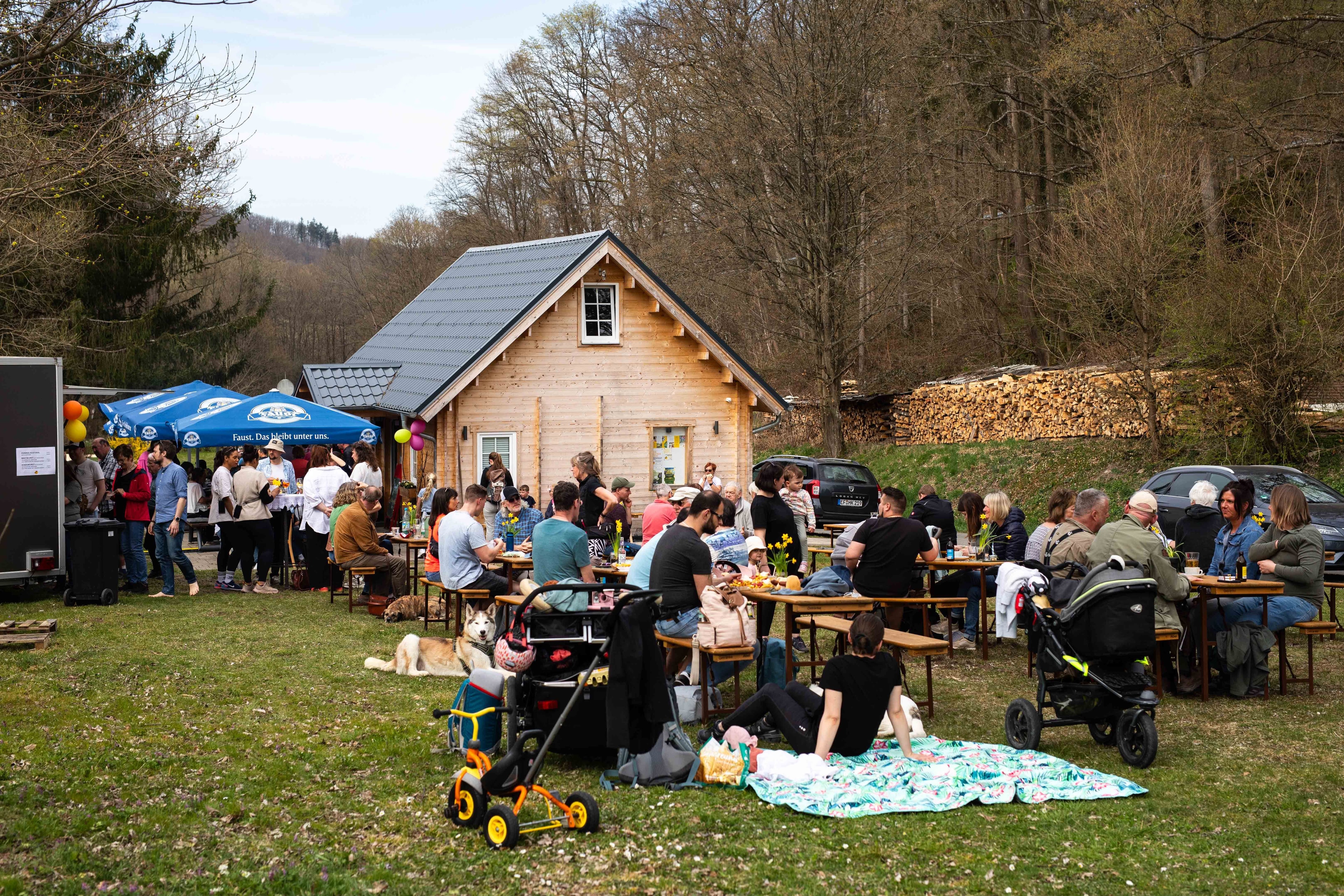 Auf einer Wiese vor einer Holzhütte sitzen und stehen viele Menschen.