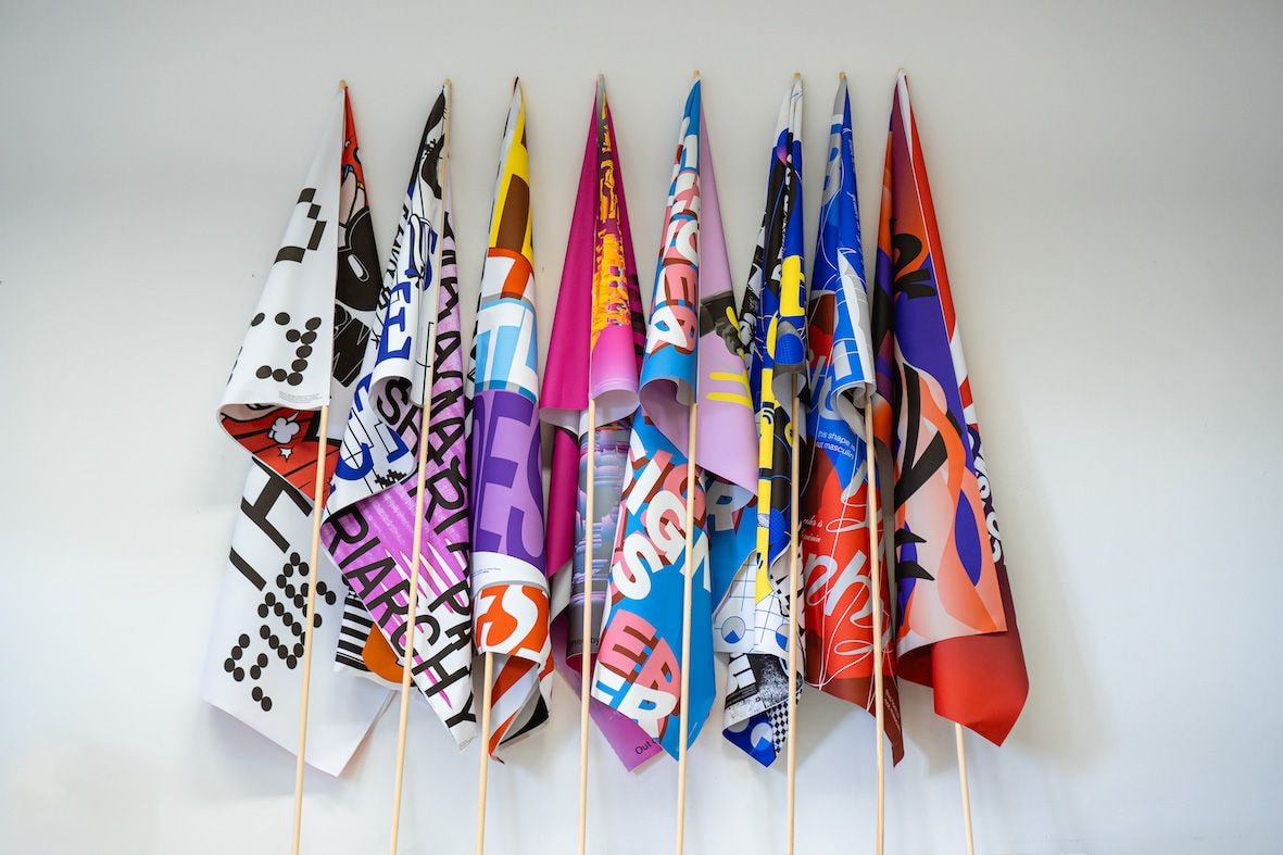 An einer weißen Wand lehnen acht bunt gemusterte Fahnen.