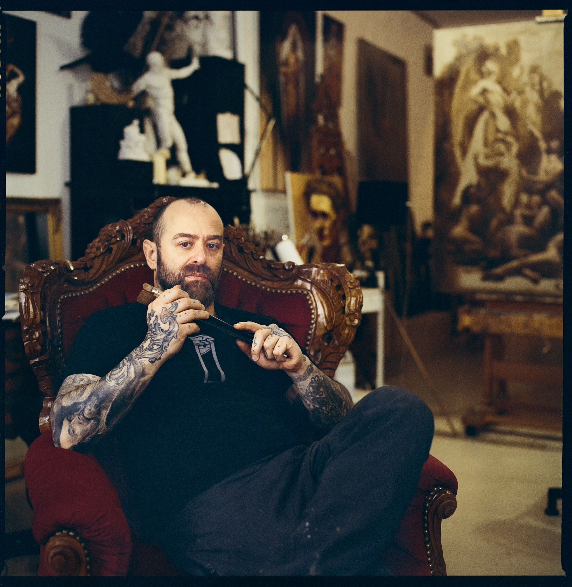 Ein tätowierter Mann sitzt in einem Kunstatelier auf einem Sessel und schaut in die Kamera.