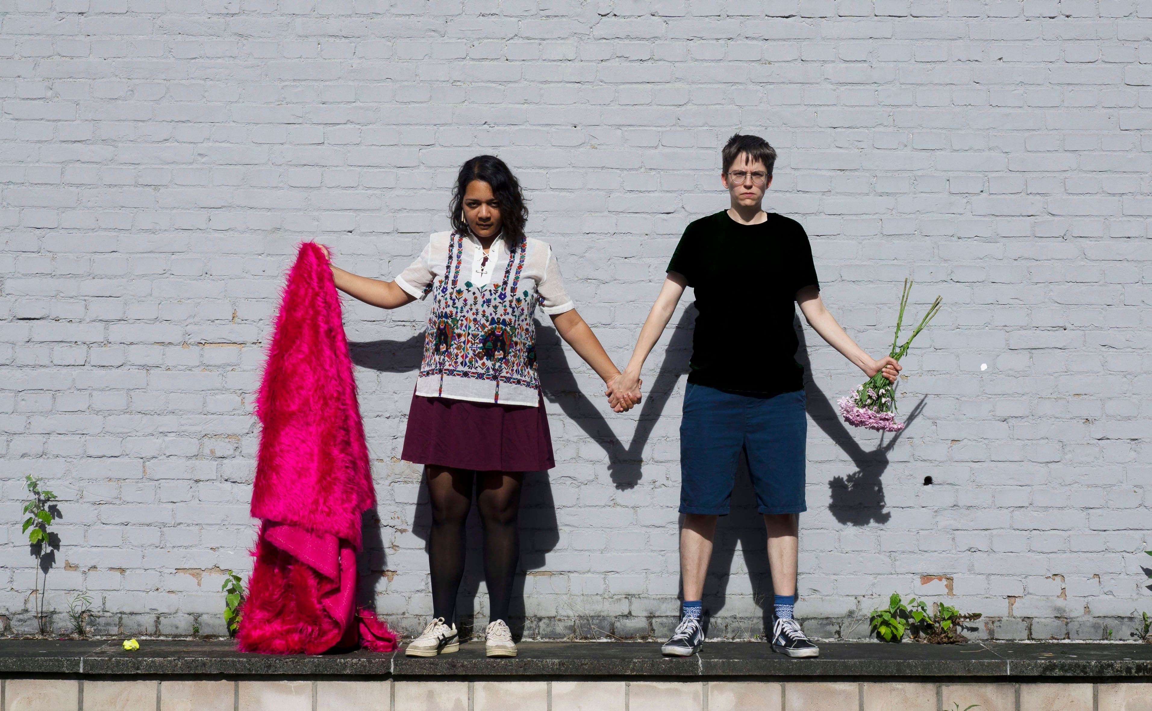Auf dem Foto stehen zwei Personen Hand in Hand auf einer Mauer und schauen streng in die Kamera. Die Person links hält eine pinke Decke in der Hand, die Person rechts einen Blumenstrauß.